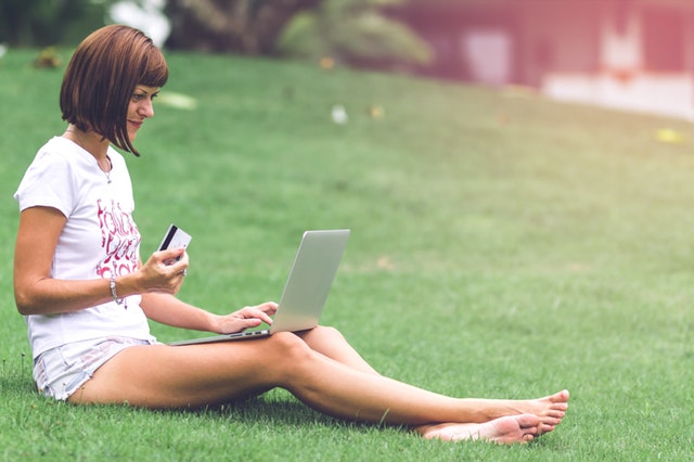 Žena s platobnou kartou v ruke sedí na trávniku a pozerá do notebooku.jpg